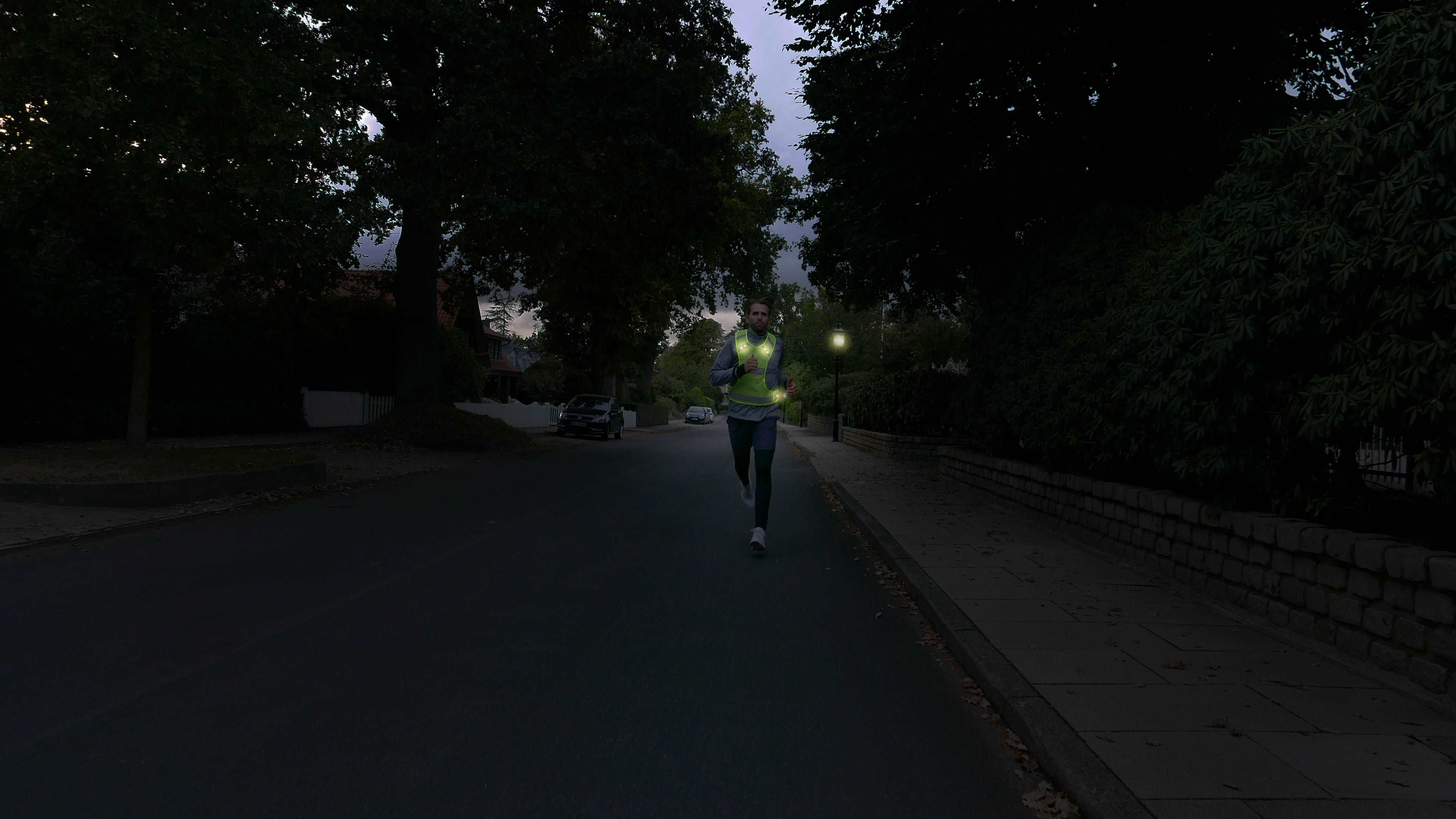 LED Sportweste mit leuchtende LEDs an joggendem Mann im Dunkeln – Straße mit Bäumen und Autos im Hintergrund 